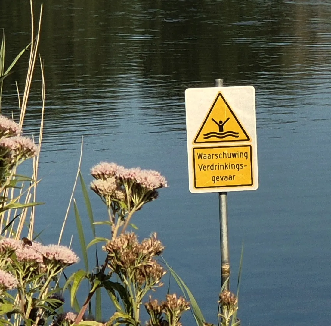 Waarschuwingsbord verdrinkingsgevaar tussen wilde bloemen bij het water